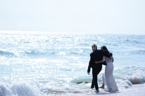 The embracing bride and groom play with their feet in the waves of the sea.