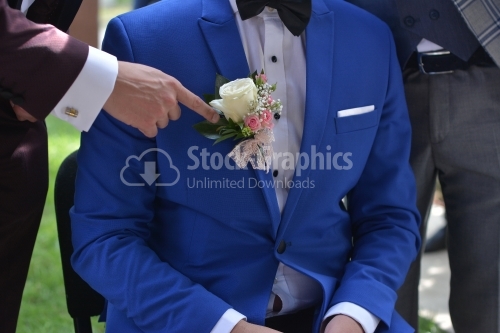 The groom sits on the chair and a man points to the flower in his chest.